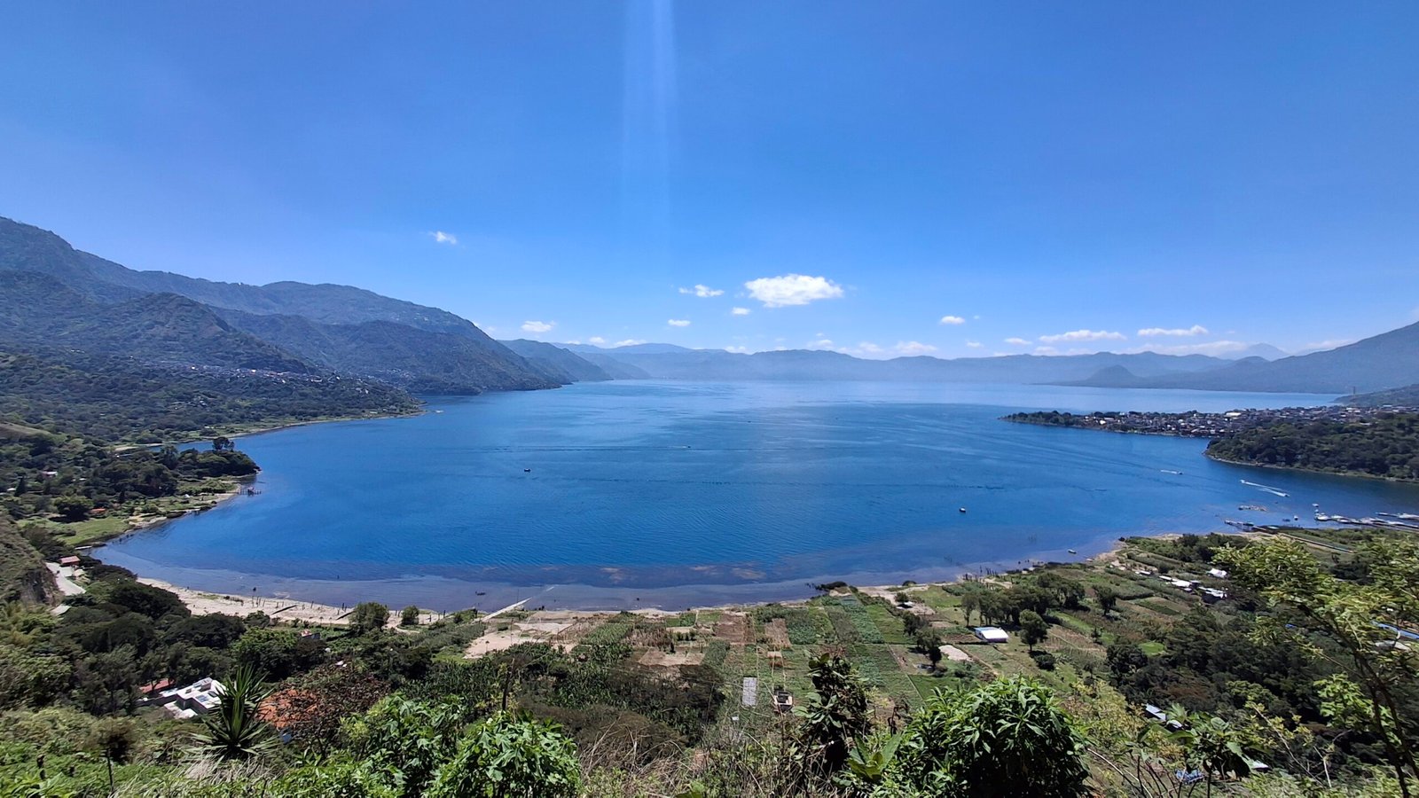 Snow-free mountain valley surrounding a calm blue lake under a clear sky; towns line the far shore.